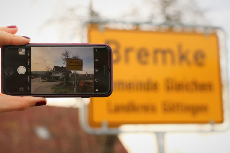Man sieht ein Smartphone, das im Foto-Modus vor das Ortsschild des Ortes Bremke gehalten wird.