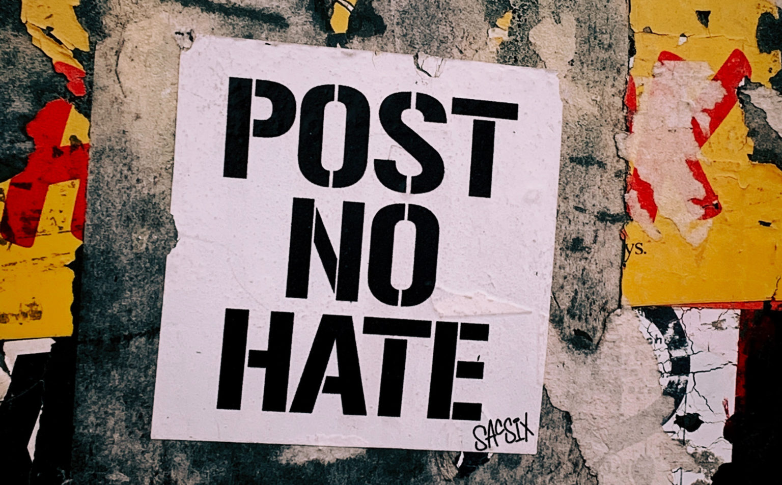 Auf einer stark abgenutzten Außenwand klebt ein Plakat in großen schwarzen Buchstaben auf weissem Grund auf dem steht: Post no hate.