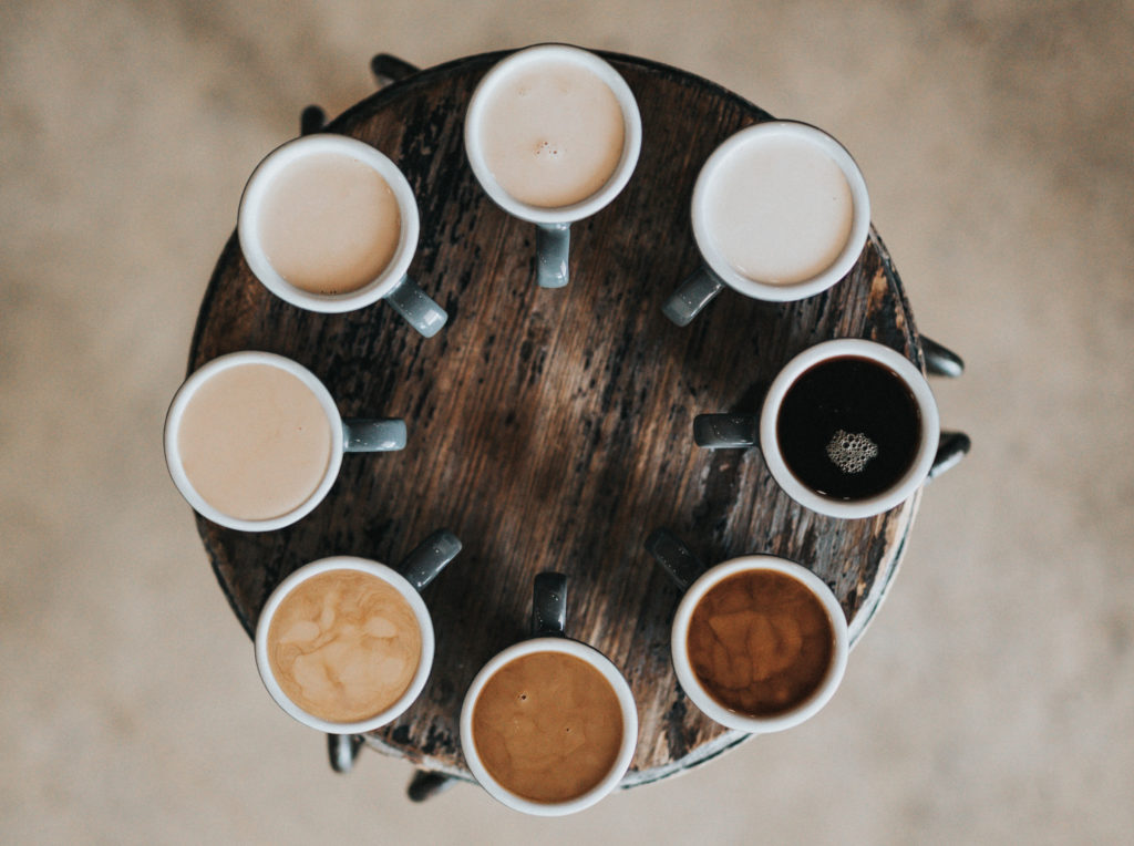 Ein runder Holztisch von oben otografiert. Auf ihm stehen 9 volle Kaffeebecher mit verschiedenen Kaffeesorten.