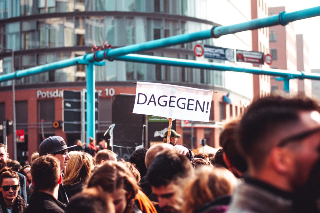 Man sieht den Ausschnitt einer Demonstration mit vielen Menschen. einer hält in der Bildmitte ein großes weisses Transparent mit der Aufschrift "Dagegen" hoch.