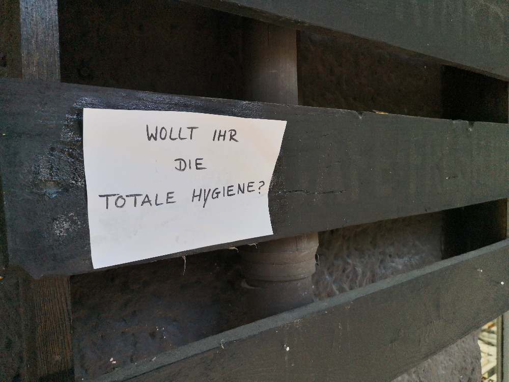 Auf einer dunkel lasierten Holzpalette an einer Fassade hängt ein handschriftlicher Zettel mit der Aufschrift "Wollt ihr die totale Hygiene?"
