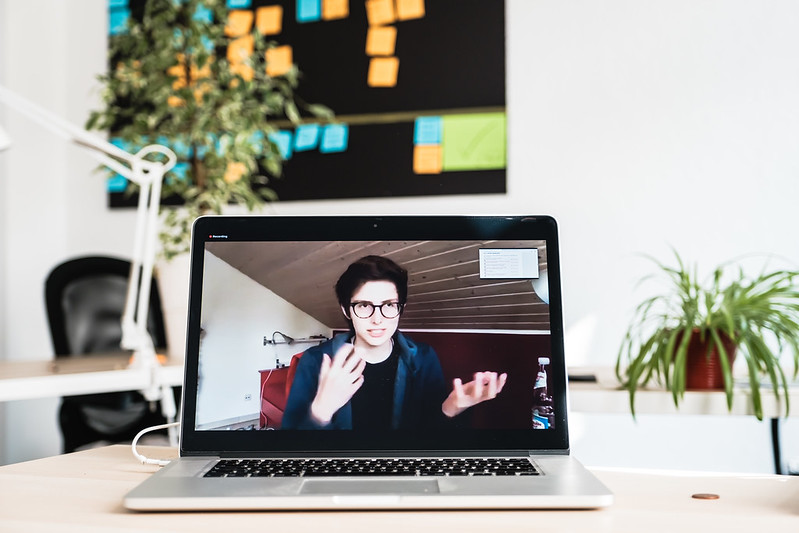 EIne Frau gestikuliert in einem Video, das auf einem Laptop abgespielt wird. Im Hintergrund sieht man ein Büro mit Pflanze und einem schwarzen Board voller Postits.