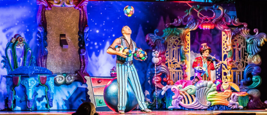 Auf einer magisch bunten und vollen Bühne jongliert ein Mann mit mehreren Bällen.