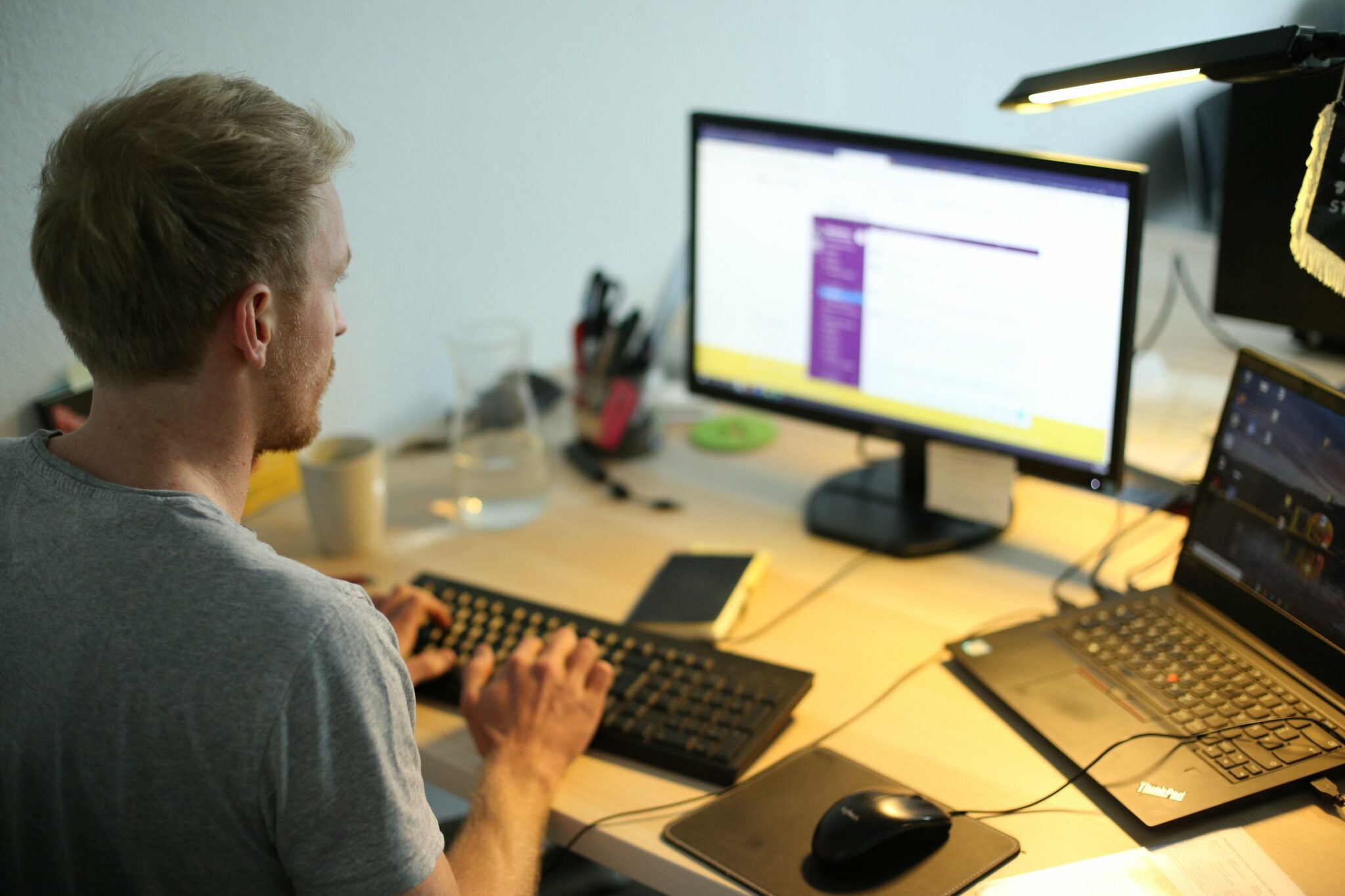 Foto zeigt einen jungen Mann im Halbprofil, der vor einem Laptop und einem zweiten Bildschirm im Schein einer Schreibtischlampe arbeitet.