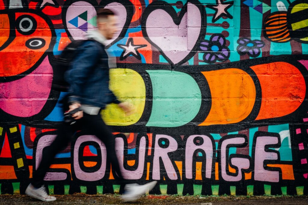 Man läuft vor einer Wand lang auf der ein Graffiti mit dem Text "Courage" steht.