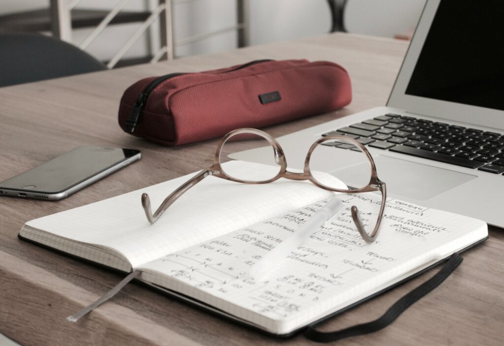 Foto eines Notizblocks, auf dem eine Brille und der vor einem Laptop liegt.