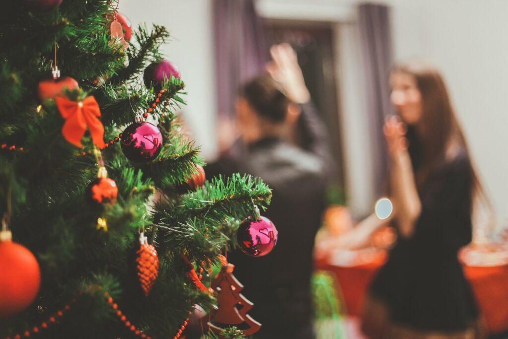 Foto einer Weihnachtsfeier. Im Vordergrund ein geschmückter Weihnachtsbaum – im Hintergrund zwei Menschen.