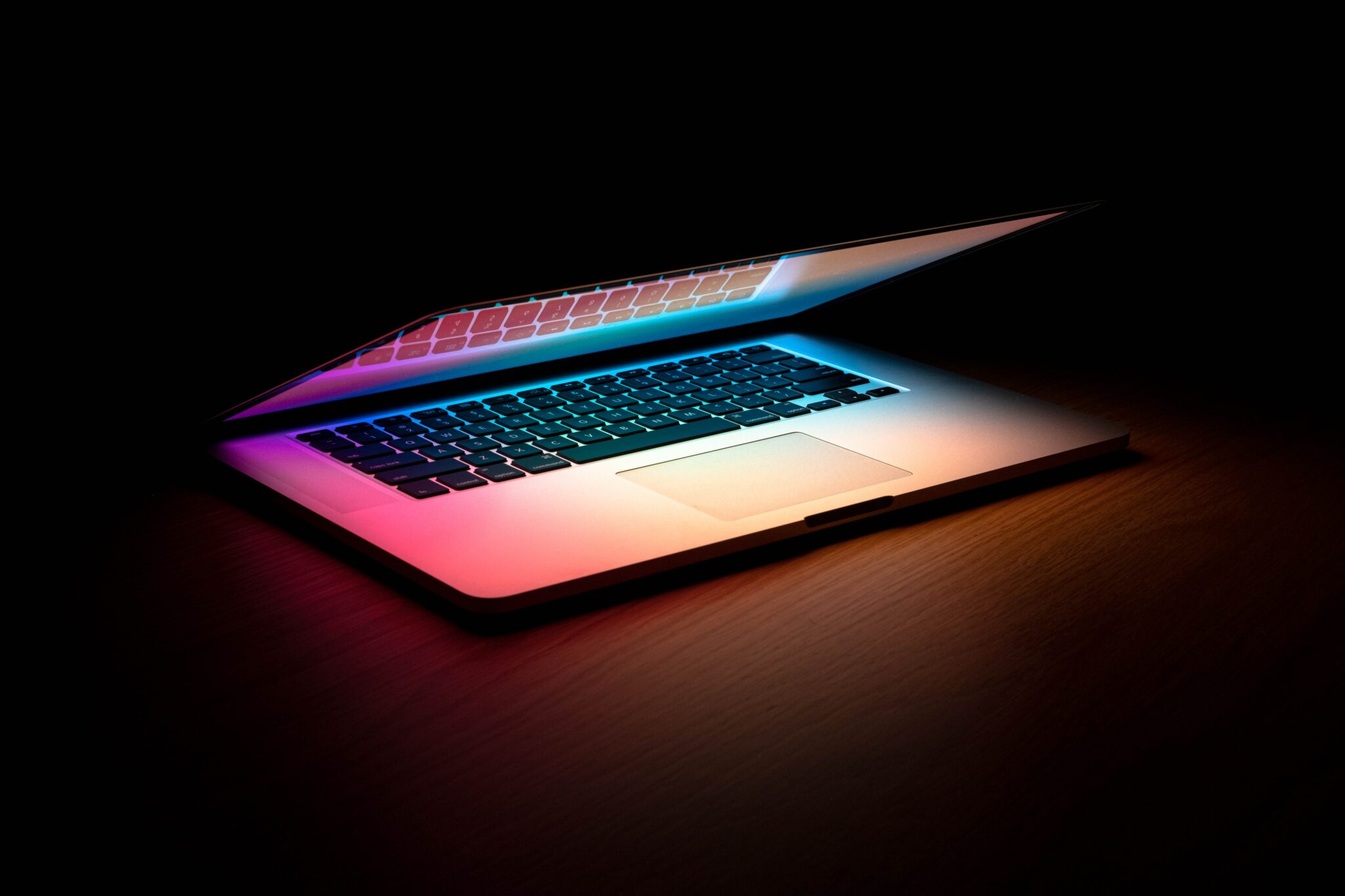 Ein Laptop steht halb zugeklappt im dunkeln, man sieht nur buntes Licht vom Bildschirm auf die Tastatur strahlen.
