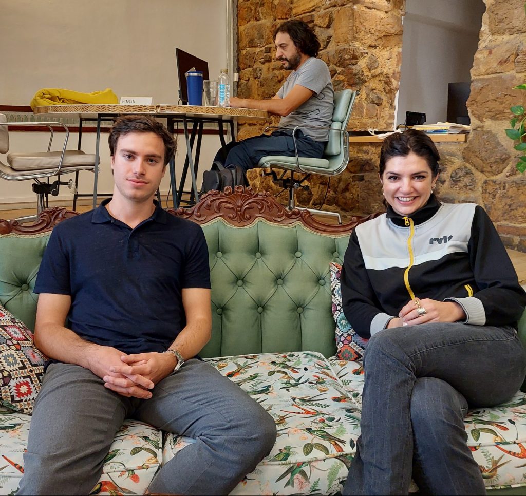 Aris und Elena sitzen auf einer grünen Couch im Impact Hub Berlin, im Hintergrund arbeitet ein Co-Worker.