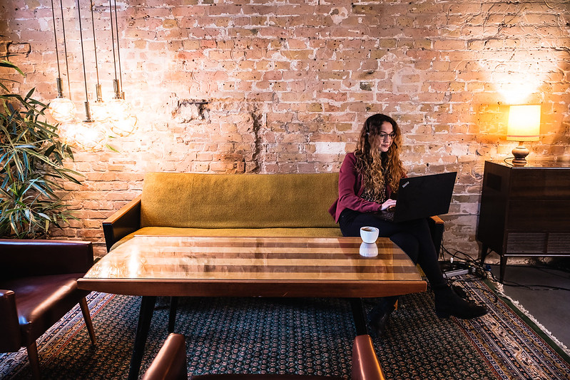 Foto: Lisa Klank sitzt allein auf einem langen Sofa an einem Tisch vor einer Backsteinwand. Sie ist in ihren Laptop vertieft.