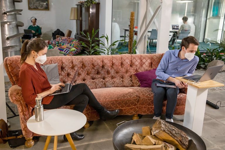 Nora Lassahn und Sebastian Gillwald sitzen auf einem langen roten Sofa, beide arbeiten an ihrem Laptop mit Jitsi, einem OpenSource Programm.