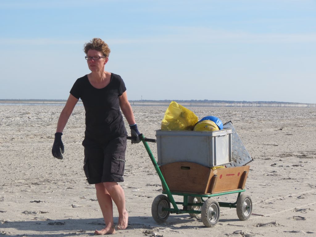 Jennifer Timmrott von Küste gegen Plastik und ReplacePlastic App sammelt mit einem Bollerwagen Müll an einem breiten Sandstrand.