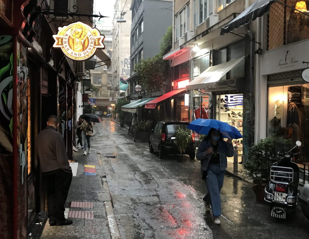 Straße in Athen bei Regen, Menschen mit Regenschirmen und spiegelnde, nasse Flächen.