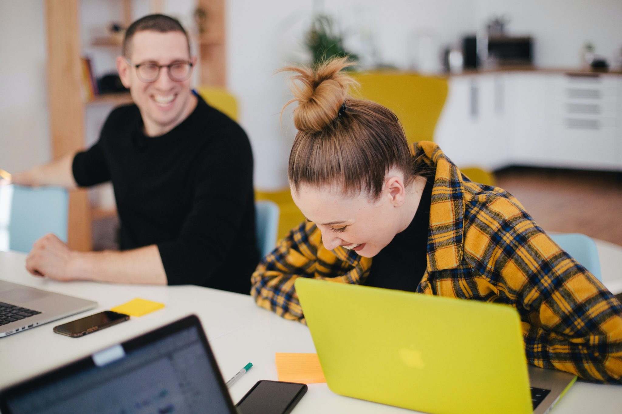 Eine junge Frau sitzt an einem Gruppentisch und lacht, vor ihr ein gelber Laptop, neben ihr im Hintergrund ein junger Mann, der sich ebenfalls amüsiert. Sie sind Teil eines digitalen Teams.