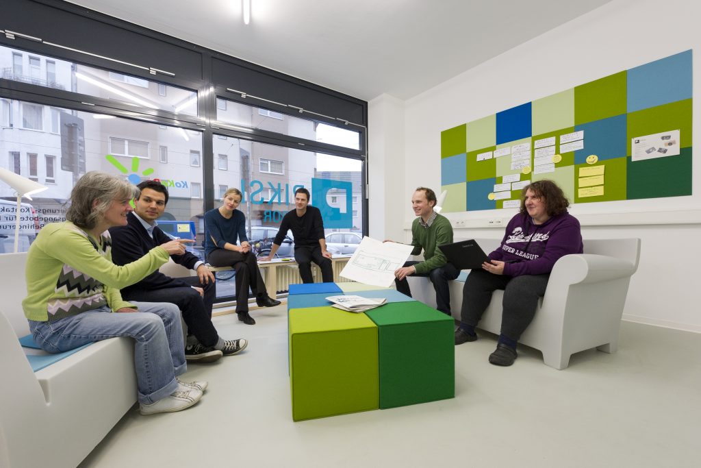 Foto zeigt eIne Gruppe Menschen mit und ohne Behinderung im PIKSL-Labor Bielefeld. Sie sitzen in einer gemütlichen Sitzecke im offenen Halbkreis auf Sofas und diskutieren.