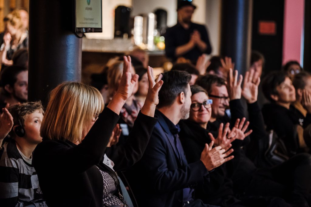Foto eines Publikums von der Seite, das Gehörlosenapplaus spendet (erhobene Hände, die wedeln).