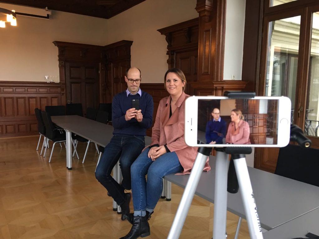 Insa Heinemann und Friedemann Schnur von der Braunschweigischen Stiftung sitzen auf einem Tisch, vor ihnen auf einem Stativ ein Handy, das sie filmt.