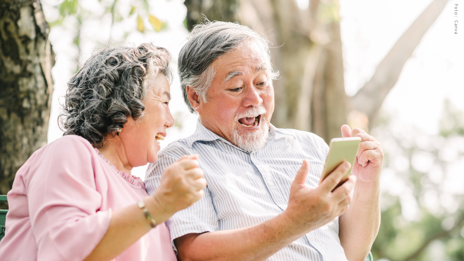 Zwei ältere Menschen sitzen im Wald auf einer Bank und blicken lachend auf ein Smartphone, das der Mann in der Hand hält.