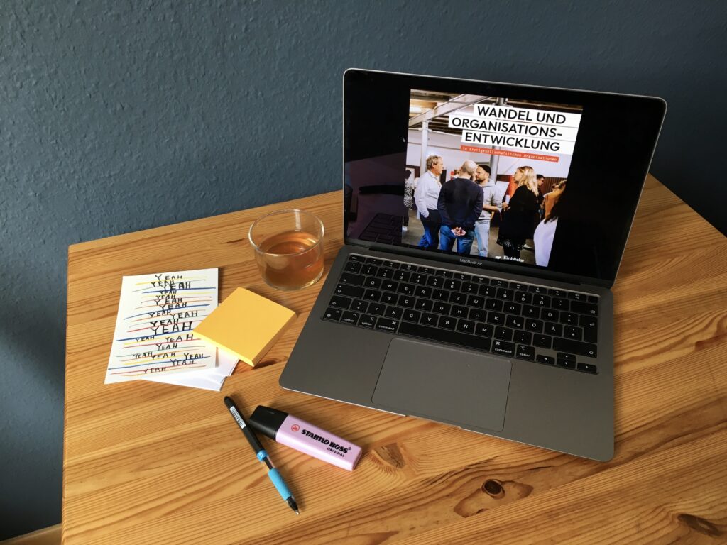 Foto eines Schreibtischs mit Stiften und Post-Its. Im Blickfeld ein Laptop, auf dem das openTransfer eBook zu Organisationsentwicklung zu sehen ist.
