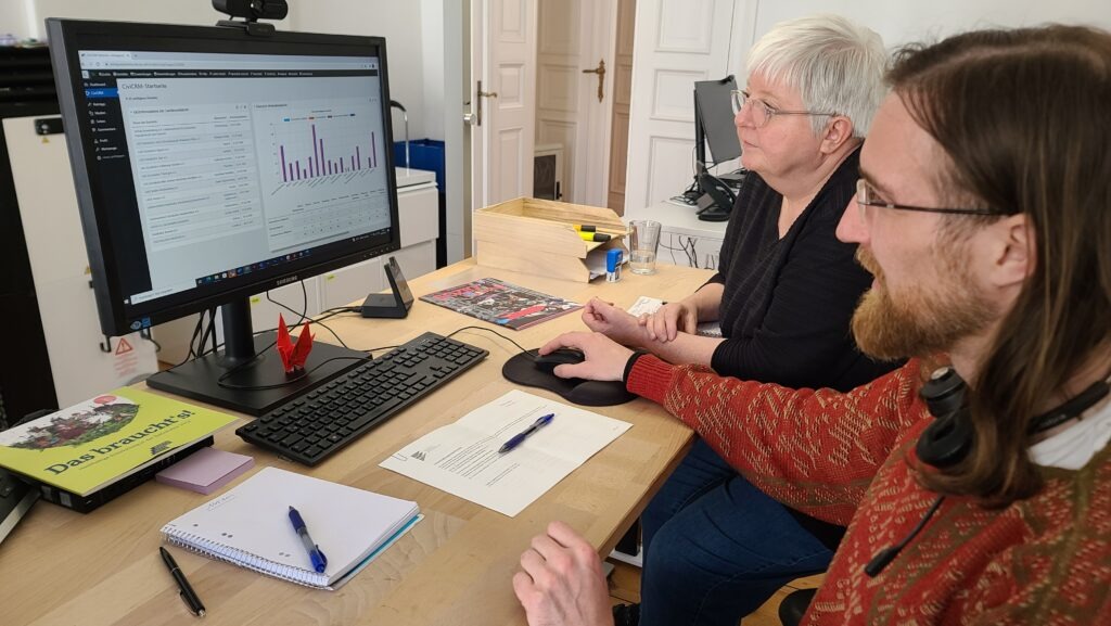Ein Mann und eine Frau sitzen an einem Schreibtisch vor einem Bildschirm, auf dem statistische Auswertungen zu sehen sind.