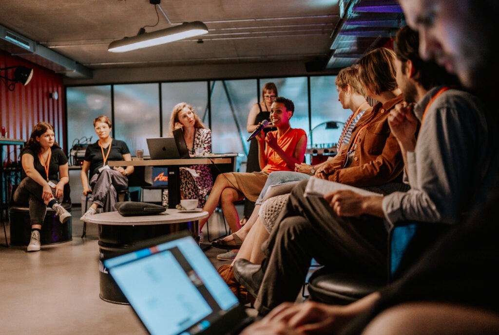 Eine Gruppe sitzt in einem Workshop-Raum, eine Person spricht ins Mikrofon, die anderen hören zu.