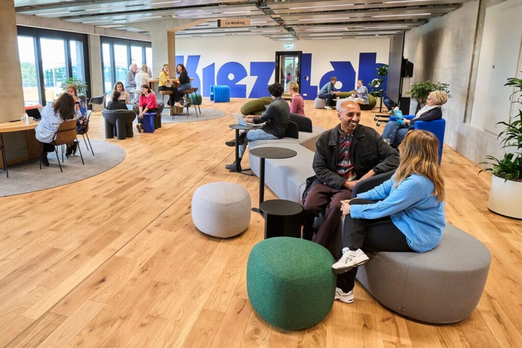 enschen sitzen und arbeiten in einem großen, hellen Co-Working-Raum des KiezLab Berlin. Der Raum ist modern eingerichtet, mit Holzfußboden, Sitzinseln und großen Fenstern