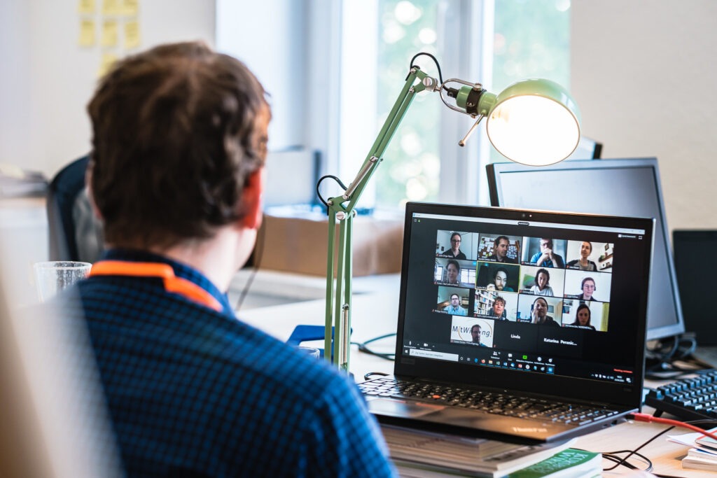 Von hinten ist ein Mann zu sehen, der auf einen Laptop schaut, auf dem eine Video-Konferenz stattfindet.