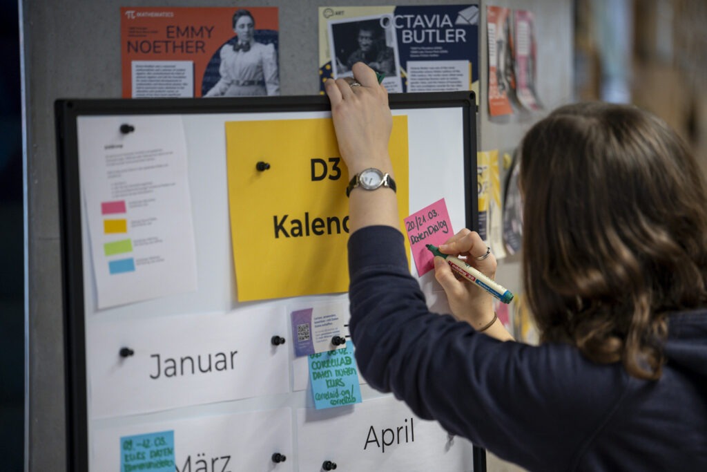 Weiblich gelesene Person schreibt Notizen auf ein Whiteboard, auf dem "D3 Kalender" steht.