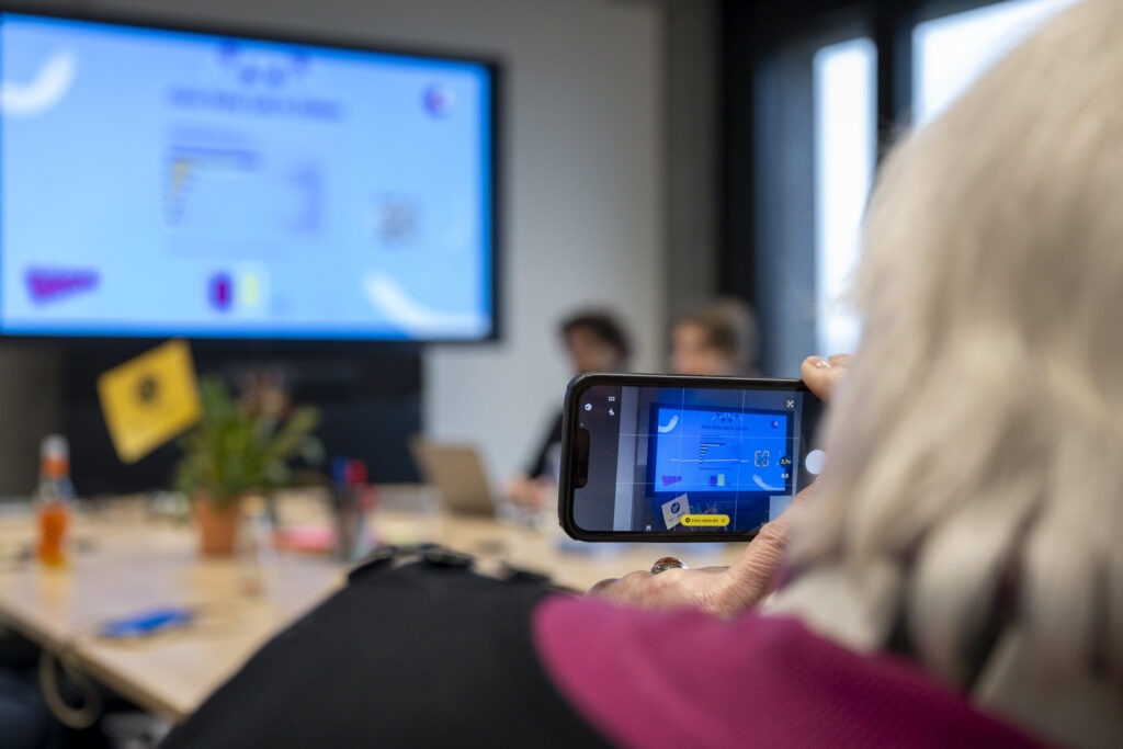 Shot über die Schulter einer anderen Person, die ein Smartphone hält und selbst ein Foto oder Video aufnimmt von einem Workshopraum.