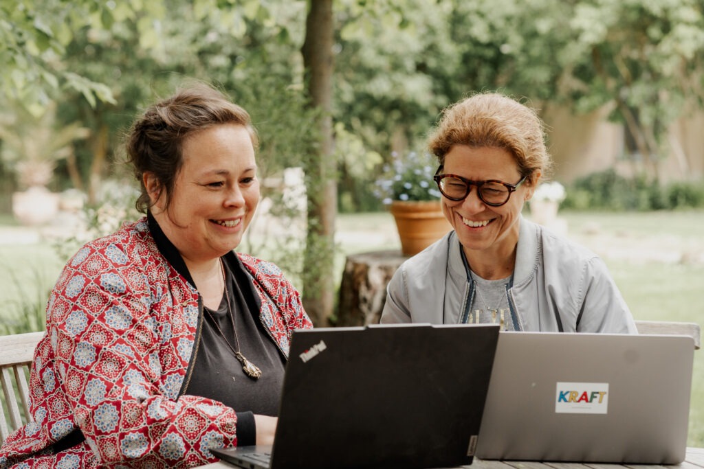 Foto von zwei weiblich gelesenen Personen jeweils vor einem Laptop an einem Tisch, miteinander redend.